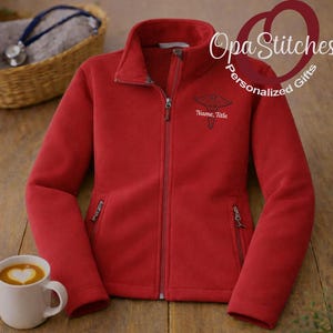 May include: A red fleece jacket with a full zipper, two side pockets, and a stand-up collar. The jacket features an embroidered medical symbol and the text "Name, Title" on the chest. A white mug with a heart-shaped design is in the lower left corner.