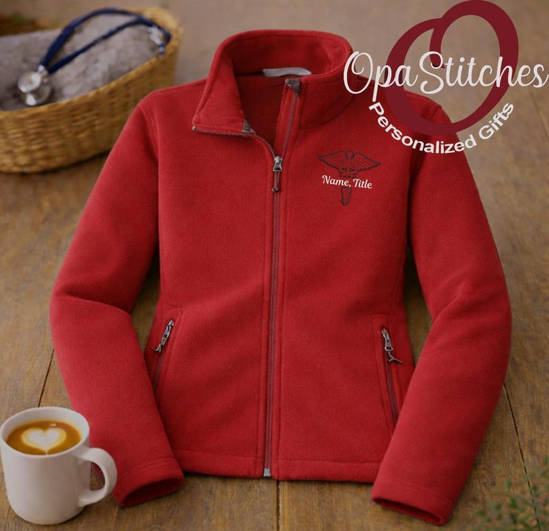 May include: A red fleece jacket with a full zipper, two side pockets, and a stand-up collar. The jacket features an embroidered medical symbol and the text "Name, Title" on the chest. A white mug with a heart-shaped design is in the lower left corner.