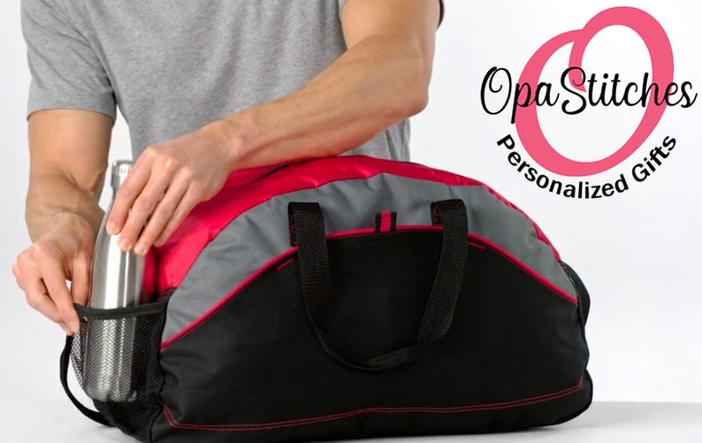 May include: A person is placing a silver water bottle into the side pocket of a black, red and grey duffel bag. The bag has a red trim and two black handles. The bag is sitting on a white surface. The text "Opa Stitches Personalized Gifts" is in the top right corner of the image.