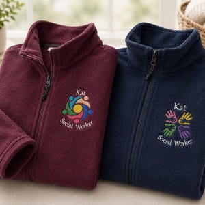 Puede incluir: Dos chaquetas polares con cremallera en color burdeos y azul marino. Cada chaqueta presenta un cuello alto y un logotipo bordado en el pecho. La chaqueta burdeos muestra un icono circular colorido con el texto Kat Social Worker. La chaqueta azul marino muestra un dise&ntilde;o de cuatro manos con el texto Kat Social Worker.