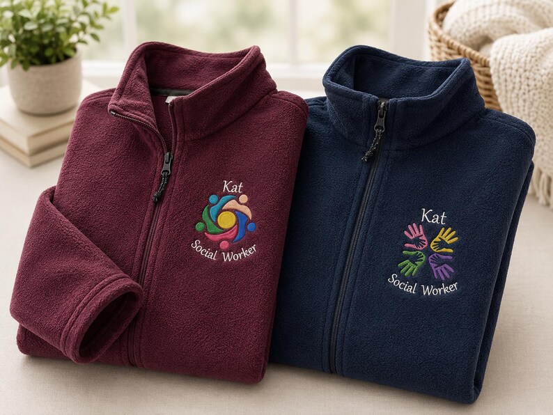 Puede incluir: Dos chaquetas polares con cremallera en color burdeos y azul marino. Cada chaqueta presenta un cuello alto y un logotipo bordado en el pecho. La chaqueta burdeos muestra un icono circular colorido con el texto Kat Social Worker. La chaqueta azul marino muestra un dise&ntilde;o de cuatro manos con el texto Kat Social Worker.