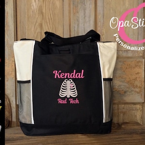 May include: Black tote bag with white side panels and mesh side pockets. The bag features the name "Kendal" and "Rad Tech" embroidered in pink, along with a rib cage graphic. The bag is designed for medical professionals.