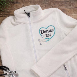 May include: A white fleece jacket with a half-zip closure. The jacket features a teal heart-shaped design with the name "Denise" and "RN" embroidered on the front, along with a stethoscope graphic. A stethoscope is visible in the foreground.