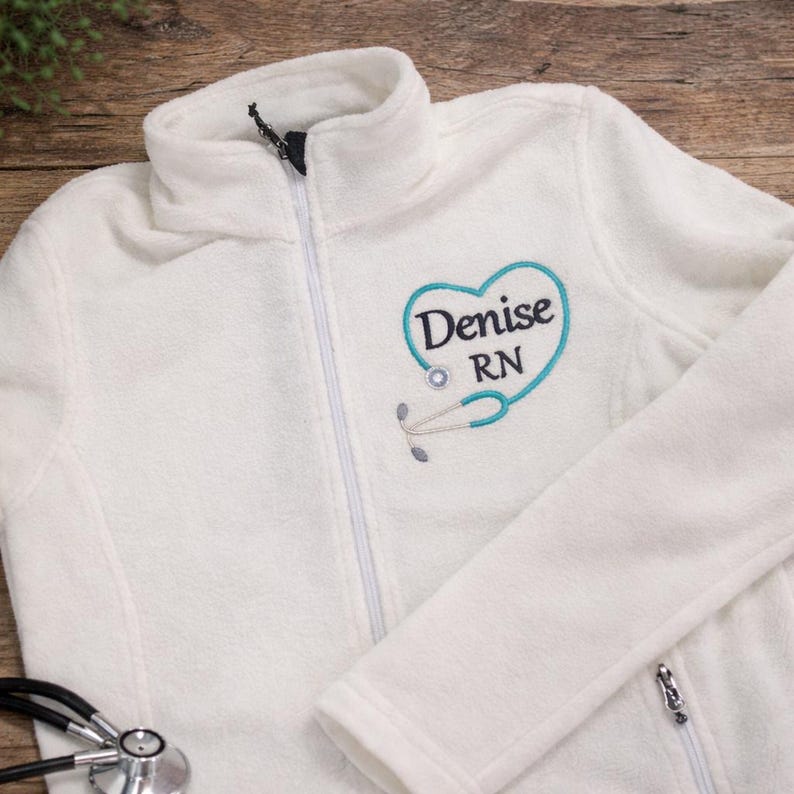 May include: A white fleece jacket with a half-zip closure. The jacket features a teal heart-shaped design with the name "Denise" and "RN" embroidered on the front, along with a stethoscope graphic. A stethoscope is visible in the foreground.