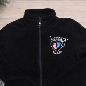 May include: Black fleece zip-up jacket with a high collar. Features a heart-shaped design with baby footprints in pink and blue, a stethoscope, and the name "Leesa" with "RN, BSN" below. A stethoscope is visible in the lower left corner.