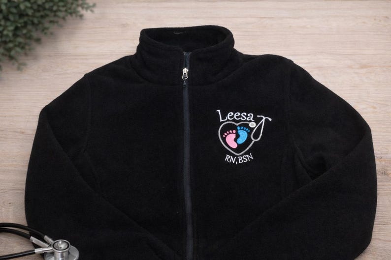 May include: Black fleece zip-up jacket with a high collar. Features a heart-shaped design with baby footprints in pink and blue, a stethoscope, and the name "Leesa" with "RN, BSN" below. A stethoscope is visible in the lower left corner.