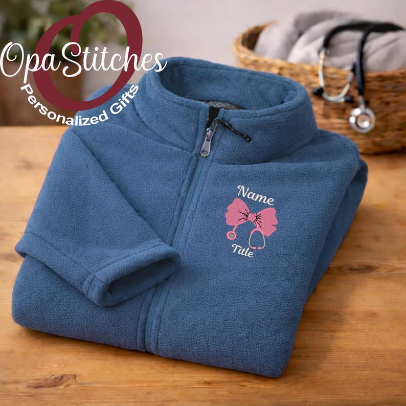 May include: A blue fleece jacket with a zipper closure. The jacket features an embroidered pink bow and stethoscope design, with the words "Name" and "Title" below. The brand name "OpaStitches Personalized Gifts" is in the upper left corner.