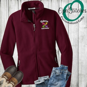May include: Burgundy fleece jacket with a zipper closure and two pockets. The jacket has an embroidered design of colorful hands with the text "Rebecca Social Worker" on the left chest.
