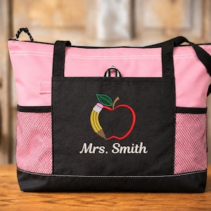 May include: A pink and black tote bag featuring a black front pocket with an embroidered design of a red apple combined with a yellow pencil. The text Mrs. Smith is embroidered in white below the apple. The bag has black straps and mesh side pockets.