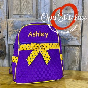 May include: Purple quilted backpack with yellow trim and a yellow polka dot bow. The name "Ashley" is embroidered in yellow thread. The backpack has a zippered front pocket and side pockets. The text "Opa Stitches Personalized Gifts" is in the background.