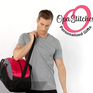 May include: A man is holding a red and black duffel bag with a grey accent. The bag has a shoulder strap. The man is wearing a grey t-shirt. The background is white. The logo " Opa Stitches Personalized Gifts" is in the top right corner of the image.