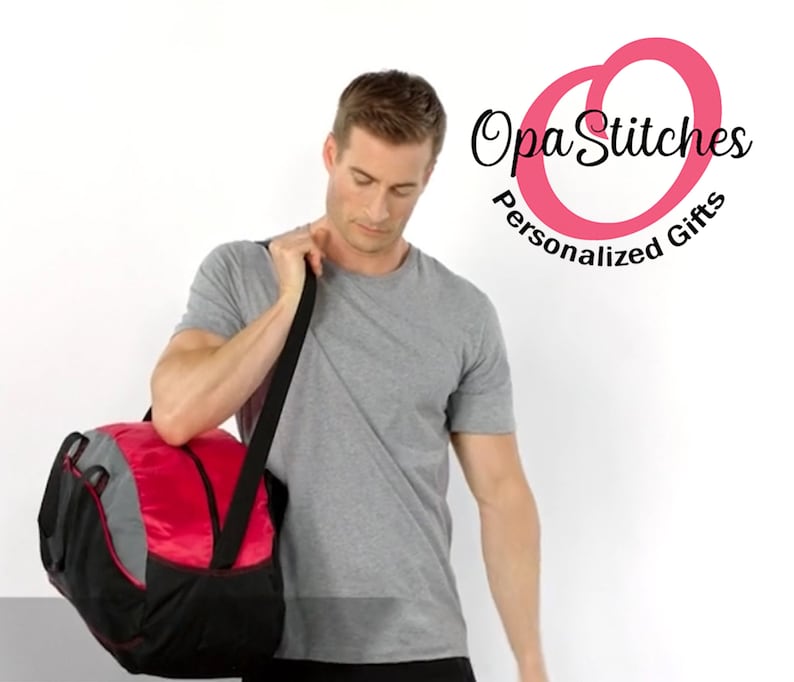 May include: A man is holding a red and black duffel bag with a grey accent. The bag has a shoulder strap. The man is wearing a grey t-shirt. The background is white. The logo " Opa Stitches Personalized Gifts" is in the top right corner of the image.