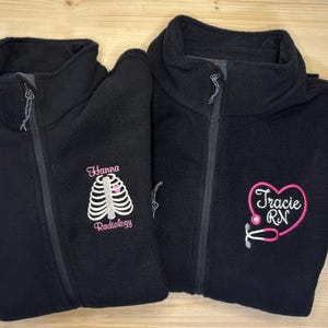 May include: Two black fleece zip-up jackets. One jacket has "Hanna Radiology" embroidered in pink with a rib cage design. The other jacket has "Tracie RN" embroidered in pink with a stethoscope design.
