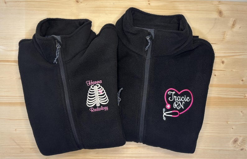 May include: Two black fleece zip-up jackets. One jacket has "Hanna Radiology" embroidered in pink with a rib cage design. The other jacket has "Tracie RN" embroidered in pink with a stethoscope design.