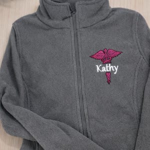 May include: A gray fleece jacket with a high collar and a full zipper. The jacket features a pink medical symbol and the name "Kathy" embroidered on the chest. A stethoscope is visible in the foreground.
