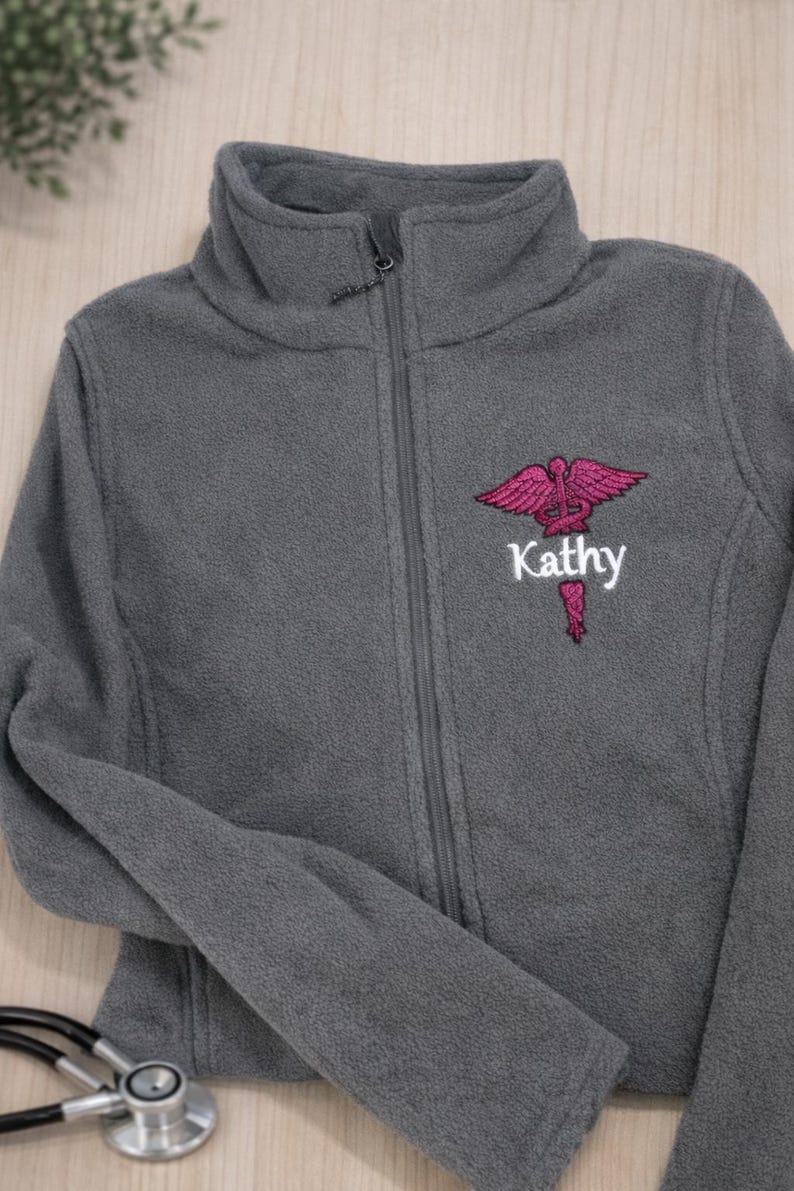 May include: A gray fleece jacket with a high collar and a full zipper. The jacket features a pink medical symbol and the name "Kathy" embroidered on the chest. A stethoscope is visible in the foreground.