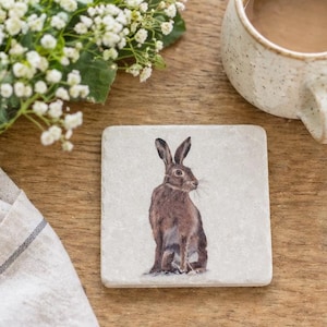 May include: Square coaster with a detailed illustration of a brown hare. The coaster has a light background and is on a wooden surface. A cup of tea and a bouquet of white flowers are also visible.