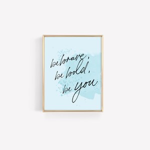May include: A light blue background with a gold frame and the text "be brave, be bold, be you" in black cursive writing.