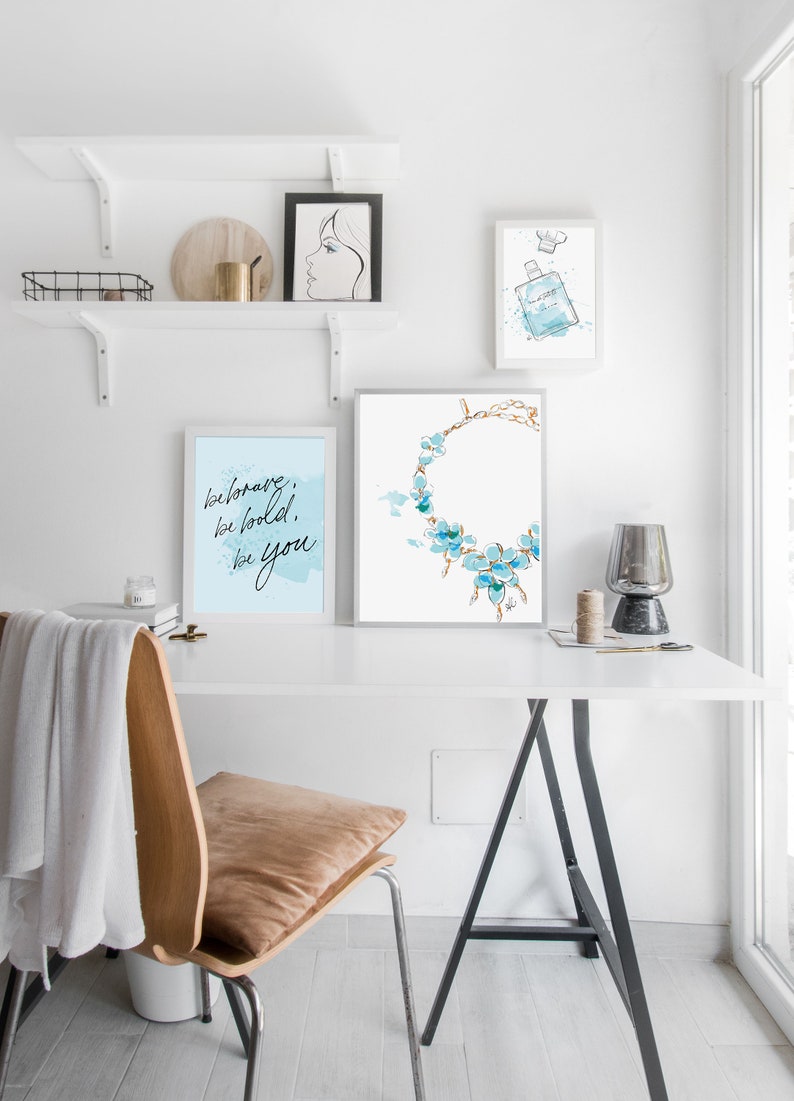 May include: A white desk with a black easel style table and a chair. There are three framed prints on the wall above the desk. The prints are of a woman's face, a bottle of perfume, and a necklace with blue flowers. The text on the print reads "be brave, be bold, be you."