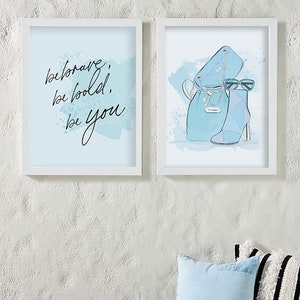 May include: Two framed prints with a blue watercolor background. The left print features the text "be brave, be bold, be you". The right print features a blue handbag, sunglasses, and a blue ankle boot.