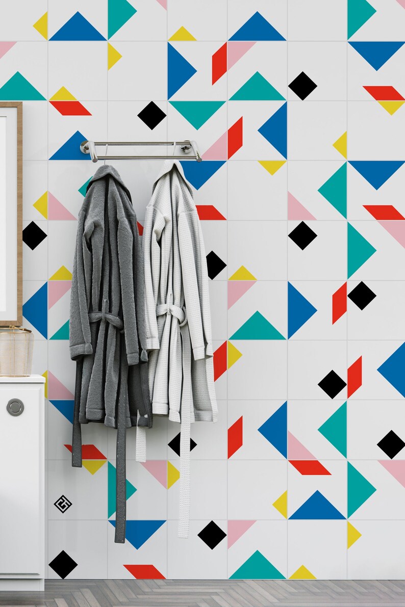 May include: Two bathrobes, one gray and one white, hang on a chrome bar against a white tiled wall. The wall features a geometric pattern of colorful shapes including triangles, diamonds, and rectangles in blue, red, yellow, and black.