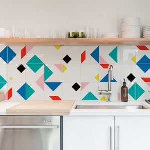 May include: Modern kitchen with white cabinets, stainless steel appliances, and a geometric tile backsplash featuring blue, red, black, pink, and yellow shapes. A wooden shelf holds glassware and plates.