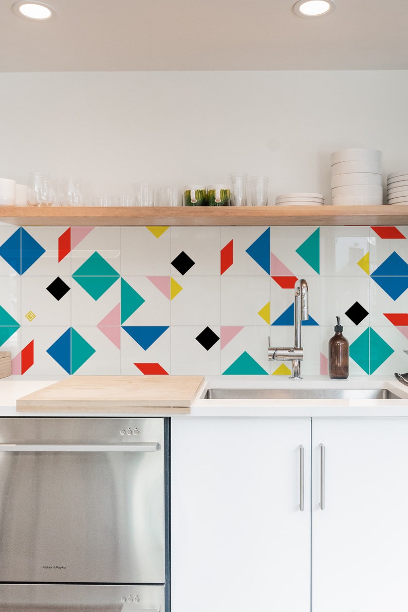 May include: Modern kitchen with white cabinets, stainless steel appliances, and a geometric tile backsplash featuring blue, red, black, pink, and yellow shapes. A wooden shelf holds glassware and plates.