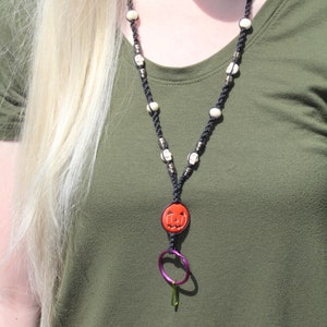Op de afbeelding: Zwarte macramé ketting met witte kralen en schedel accenten. De hanger is een ronde oranje pompoen met een uitgesneden gezicht. Een paarse ring en groene clip zijn aan de hanger bevestigd.