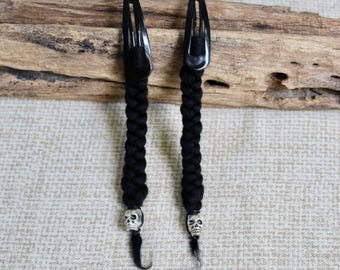 May include: Two black hair clips featuring a braided black cord attachment. Each clip ends with a small, metallic silver-colored skull bead and a short, loose strand of black hair extending from the bottom of the skull charm.