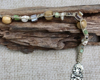 May include: A knotted beige cord bracelet featuring a variety of beads including smooth tan wooden spheres, small green stone chips, white skull-shaped beads, metallic silver cylinders, and a silver-toned skull and crossbones charm pendant.