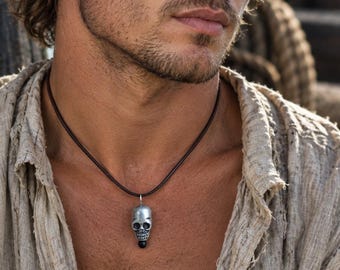 May include: A silver-toned skull pendant suspended from a dark brown leather cord necklace. The pendant features a detailed human skull design with a small black bead accent at the base, worn around the neck against a light beige textured shirt.