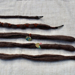 Può includere: Sei dreadlock marroni con diverse perline e ciondoli decorativi. Le dreadlock sono disposte in fila su uno sfondo di tessuto bianco.
