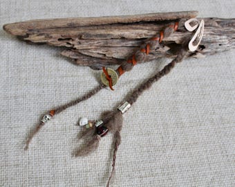 May include: Two brown hair extensions made of wool, featuring a braided design wrapped with thin leather cord. The extensions are adorned with a round brass coin, a silver-toned skull bead, a dark red bead, and a small white skull charm.