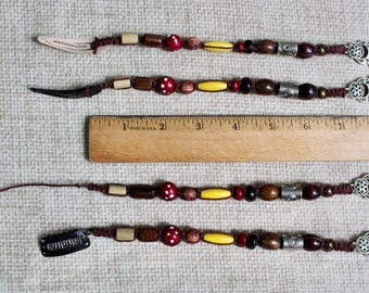 May include: Four hair extensions featuring brown, red, yellow, and black beads, including red beads with white dots, silver-toned metal accents, and silver-toned charms at the ends. A wooden ruler shows the items measure approximately 8 inches long.