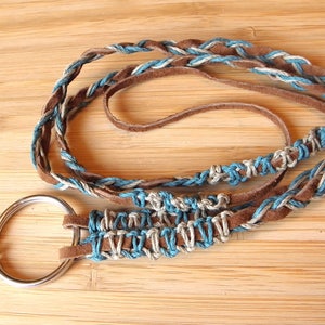 May include: A braided leather lanyard with a silver ring and a clip. The lanyard is made of brown leather, blue cord, and white cord.
