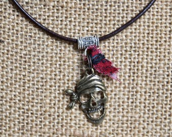 May include: A dark brown leather cord necklace featuring a metallic silver-toned bail and a bronze-toned skull pendant wearing a bandana. A small piece of frayed red and black plaid fabric is attached to the bail, hanging beside the skull charm.