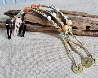 May include: Three hair extensions featuring metal snap clips in black and beige, attached to braided cords adorned with assorted wooden, glass, and shell beads in shades of green, white, and tan, finished with round brass-toned metal coins.