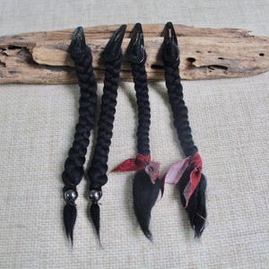Puede incluir: Cuatro pinzas para el cabello negras con extensiones trenzadas. Dos pinzas tienen una pequeña cuenta plateada en la base de la trenza, mientras que las otras dos presentan un detalle de tela con estampado rojo y negro atado en la parte inferior.