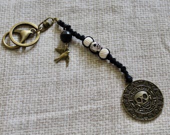 May include: A keychain featuring a bronze-toned metal ring and clasp attached to a black cord. The cord is adorned with a black bead, a small bronze bird charm, two white beads, and a silver-toned skull bead. A large circular bronze coin pendant with a skull design hangs at the end.