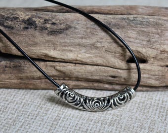May include: A black cord necklace featuring a curved, silver-toned metal tube bead. The bead has a repeating embossed spiral pattern across its surface. The cord is smooth and thin, passing through the center of the cylindrical metal component.