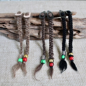 May include: Six hair extensions attached to metal snap clips, featuring braided strands in light brown, dark brown, and black. Each braid is decorated at the bottom with metallic, green, red, and gold-colored beads, ending in loose hair fibers.