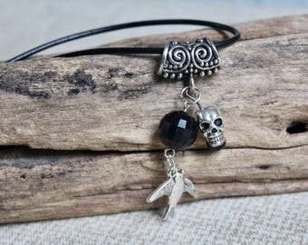 May include: A black cord necklace featuring a silver-toned metal bail with a spiral pattern. Suspended from the bail are three charms: a faceted black bead, a small silver-toned metal skull, and a silver-toned metal bird in flight.