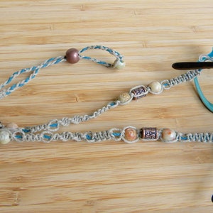 May include: A glasses chain crafted from braided beige and blue cord, featuring a sliding brown bead for adjustment. The cord is accented with various round and cylindrical beads in metallic, cream, and marbled tones, attached to turquoise eyeglass frames.