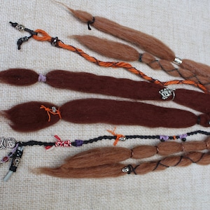 May include: A collection of brown and auburn dreadlocks, adorned with decorative beads, silver rings, and orange and black accents. The dreadlocks vary in length and thickness, with some braided and others left loose.