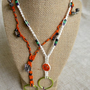 Op de afbeelding: Twee handgemaakte macramé sleutelhangers op een jute oppervlak. Eén sleutelhanger is oranje en zwart, met een zilveren bedel en groene ring. De andere is wit, met een vredesteken en groene ring. Beide hebben decoratieve kralen.
