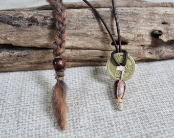 May include: A brown braided hair extension with a dark wooden bead and a loose tuft at the end, paired with a dark leather cord necklace featuring a circular brass-colored metal coin pendant with a square center hole and a small bead accent.