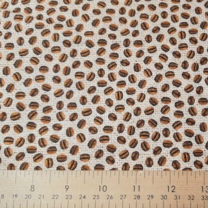 May include: Fabric featuring a repeating pattern of brown coffee beans on a beige background. A wooden ruler with centimetre markings is at the bottom of the image, measuring from 33 to 96 cm.