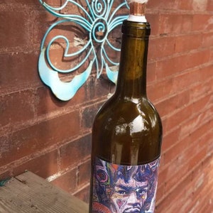May include: A brown glass wine bottle with a colorful label featuring a psychedelic portrait of a person. The bottle has a copper top with a white cotton stopper.