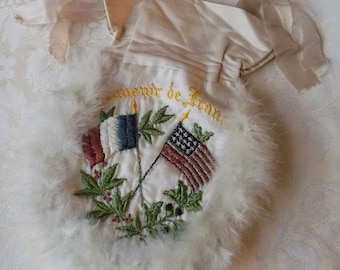 Könnte beinhalten: Eine weiße Satintüte mit einem flauschigen weißen Besatz. Die Tasche ist mit dem Text "Souvenir de France" und zwei gekreuzten Flaggen bestickt, einer amerikanischen und einer französischen.