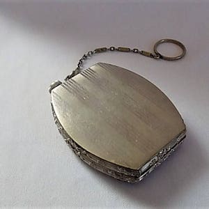 K&ouml;nnte beinhalten: Ein silbernes Kompakt mit einer Kette und einem Ring. Das Kompakt hat eine strukturierte Oberfl&auml;che und ein dekoratives Design an den R&auml;ndern.